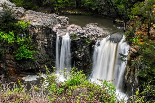 A serene waterfall cascading over rocky cliffs into a calm river, surrounded by lush green foliage.