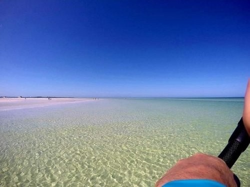 A clear, tranquil beach with shallow water and a blue sky, featuring a person's hand holding a paddle in the foreground.