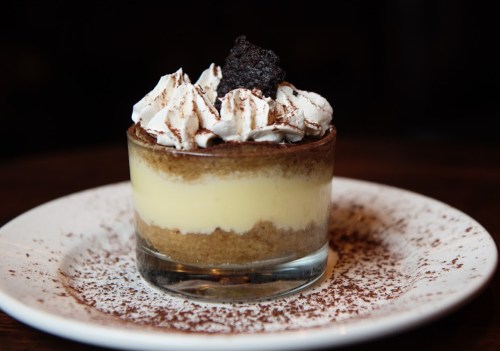 A layered dessert in a glass with creamy filling, topped with whipped cream and a chocolate cookie, on a plate with cocoa dust.