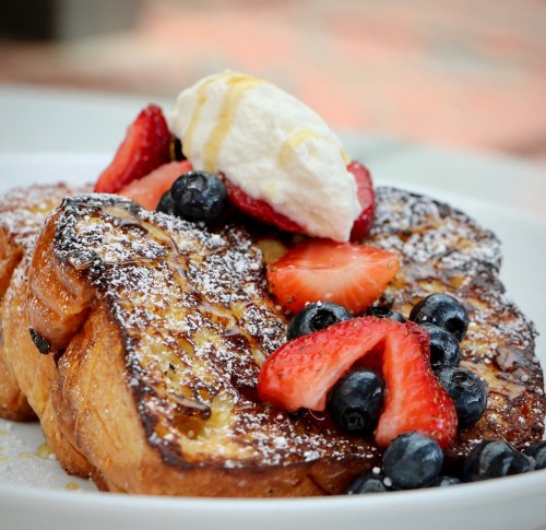 A plate of French toast topped with strawberries, blueberries, whipped cream, and a drizzle of syrup.