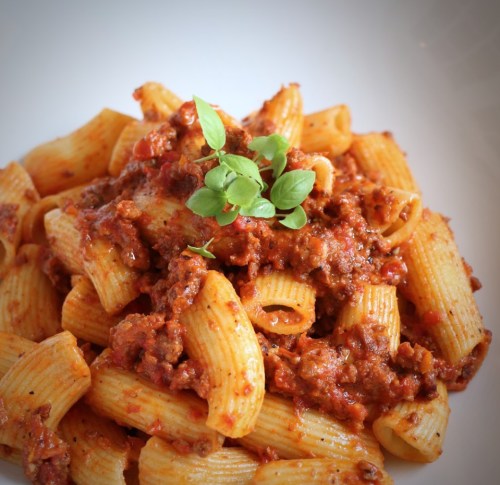 A plate of rigatoni pasta topped with a rich meat sauce and garnished with fresh basil.