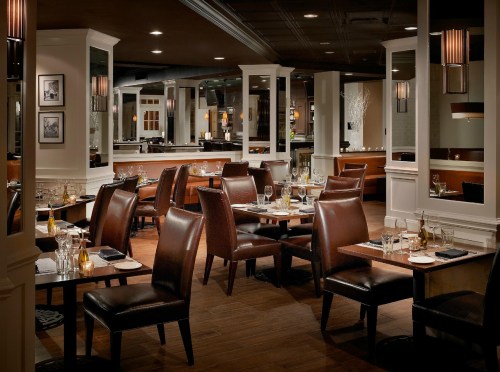 Elegant restaurant interior with wooden floors, brown leather chairs, and tables set with glassware and candles.