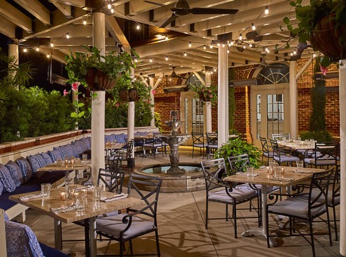 Cozy outdoor restaurant patio with string lights, potted plants, and a central fountain, featuring tables and chairs.