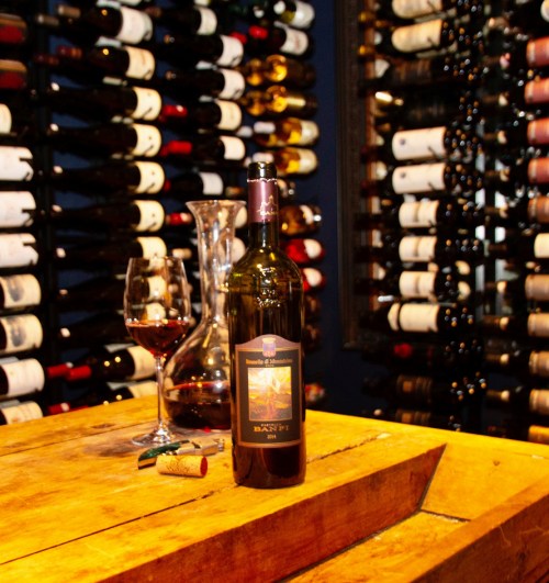 A bottle of red wine on a wooden table, with a glass and decanter, surrounded by a wall of wine bottles.