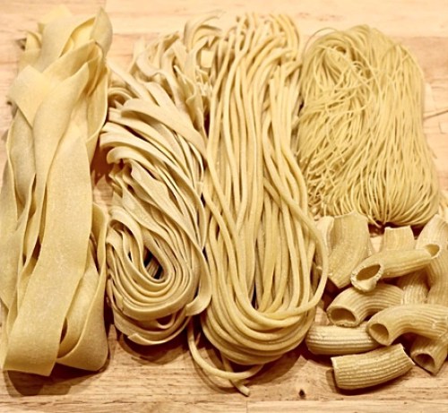 Various types of uncooked pasta arranged on a wooden surface, including fettuccine, spaghetti, and rigatoni.