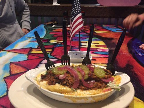 A colorful dish topped with pulled meat, pickles, and onions, served with plastic forks and a small American flag.