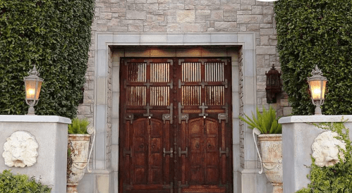 A grand entrance with double wooden doors, flanked by potted plants and stone walls, illuminated by lanterns.