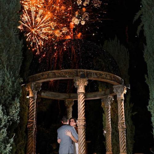 A couple embraces under a decorative gazebo as colorful fireworks light up the night sky.