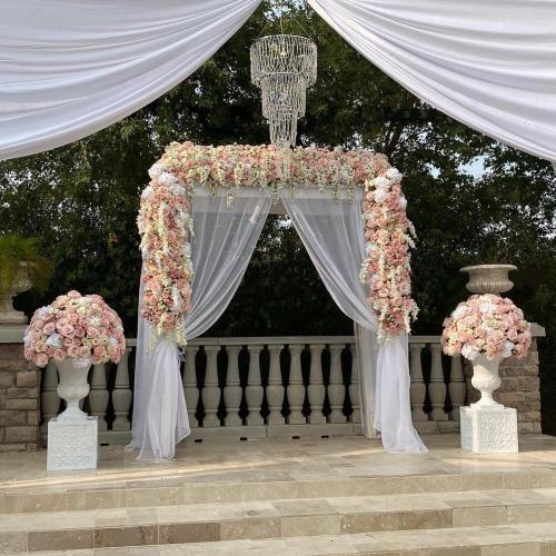 A beautifully decorated wedding arch with pink flowers, draped fabric, and a chandelier, set against a scenic backdrop.