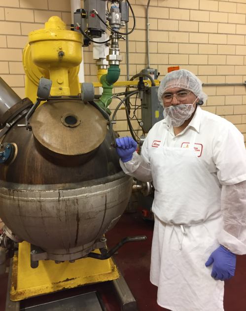 A worker in a hairnet and white coat stands next to a large industrial mixer in a food processing facility.