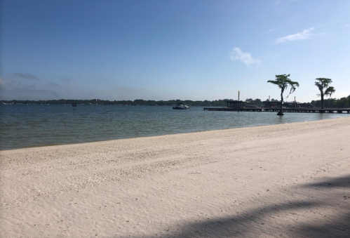 A serene beach scene with soft sand, calm water, and a distant pier under a clear blue sky.
