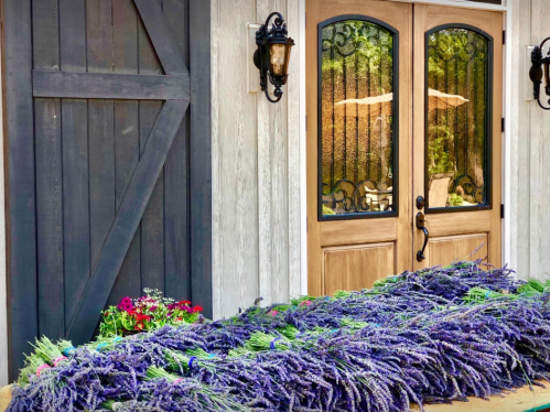 A rustic entrance with wooden doors, flanked by lavender bundles and colorful flowers on the ground.