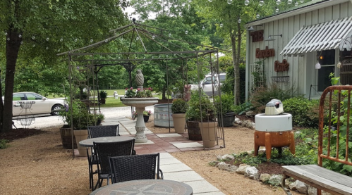 A charming outdoor café area with a gazebo, potted plants, and tables, surrounded by greenery and string lights.