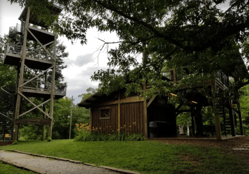A wooden building with a tower nearby, surrounded by trees and flowers under a cloudy sky.