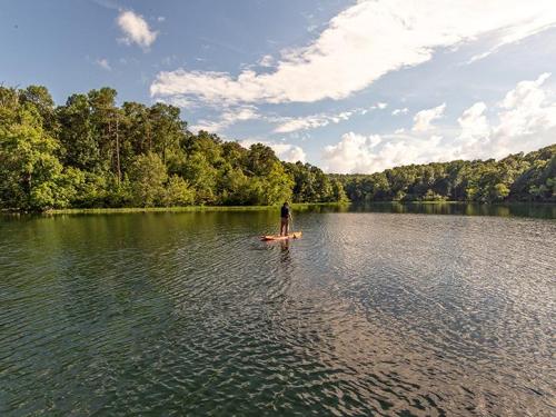 A person stands on a paddleboard in a calm lake surrounded by lush green trees and a blue sky with clouds.