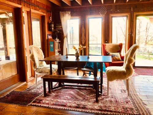 Cozy dining area with a wooden table, two chairs, a bench, and large windows overlooking a natural landscape.