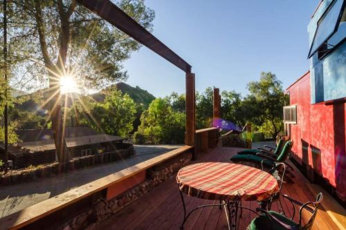Sunlight streams through trees onto a wooden deck with a round table and lounge chairs, overlooking a scenic landscape.