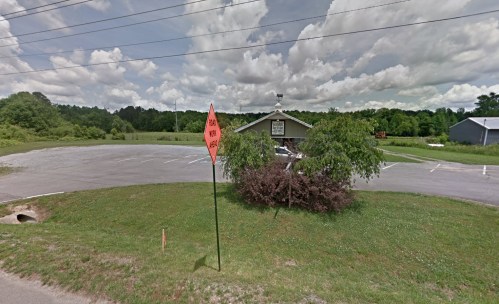 A vacant parking lot with a building in the background, surrounded by greenery and a warning sign in the foreground.