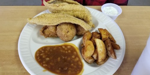 A plate of fried catfish, hushpuppies, baked beans, and potato wedges, with a small cup of dipping sauce.