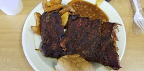 A plate of barbecue ribs with seasoned potato wedges, baked beans, and a soft bun on the side.