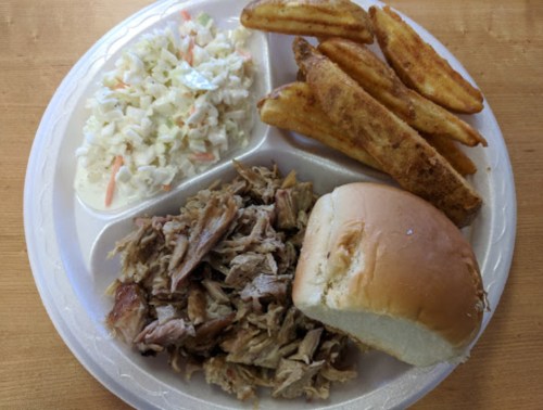 A plate with pulled pork, a bun, coleslaw, and crispy potato wedges.