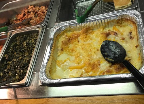 A serving tray with baked macaroni and cheese, alongside pulled pork and collard greens in a cafeteria setting.