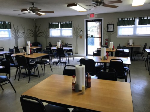 A spacious dining area with wooden tables, black chairs, and condiments on each table, featuring ceiling fans and windows.
