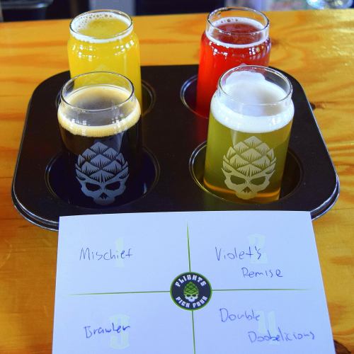 Four small glasses of beer in a flight tray, labeled Mischief, Violet's Premise, Brawler, and Double Dobleicious.