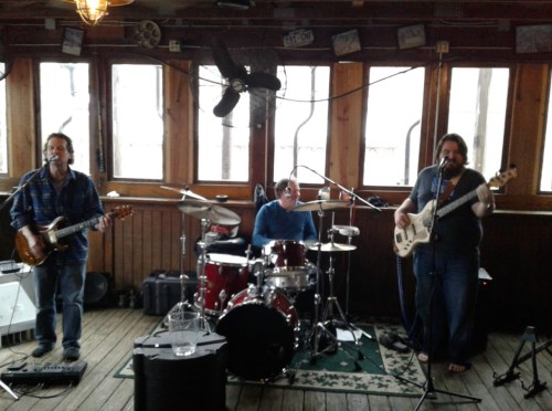 A band performs indoors with a guitar, drums, and bass, surrounded by large windows and wooden decor.