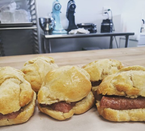 Four freshly baked sausage rolls on a wooden surface, with kitchen appliances visible in the background.