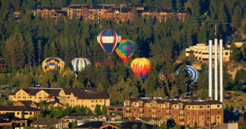 Colorful hot air balloons float above a tree-lined area with buildings in the foreground and mountains in the background.