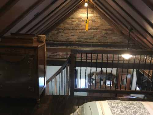 View from a lofted bedroom showing exposed brick walls, wooden beams, and a staircase leading down.