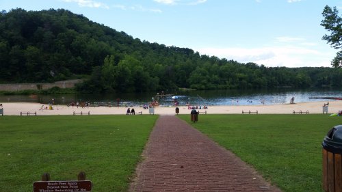 A sandy beach by a lake surrounded by green trees, with people swimming and relaxing on a sunny day.