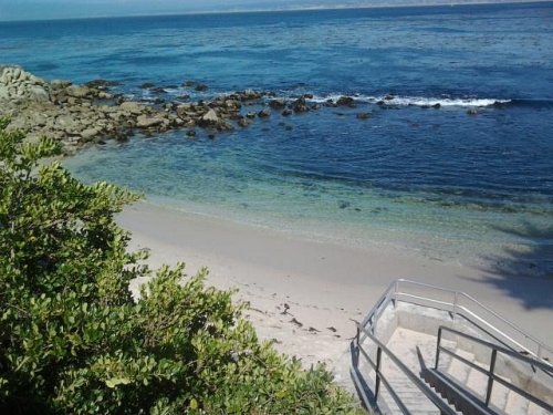 A serene beach scene with clear blue water, rocky shoreline, and a staircase leading down to the sand.