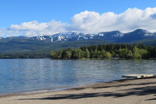 A serene lake surrounded by lush trees and snow-capped mountains under a blue sky with fluffy clouds.