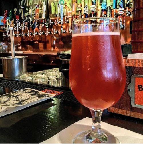 A glass of amber beer sits on a bar counter, with a row of colorful beer taps in the background.