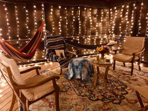Cozy outdoor space with hammocks, string lights, chairs, a table, and a floral arrangement on a patterned rug.
