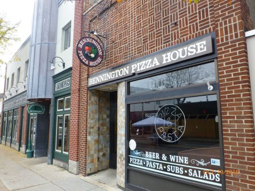 Exterior of Bennington Pizza House, featuring a sign with menu items and nearby restaurants in a brick building.
