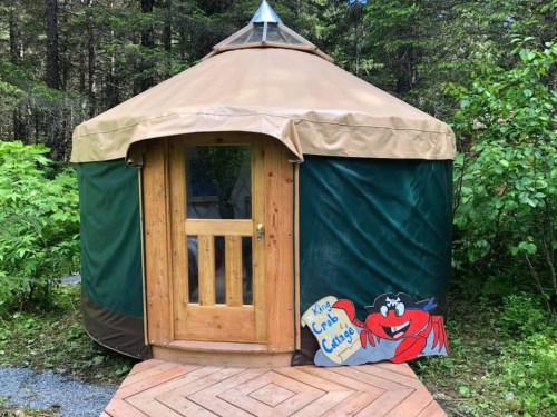 A cozy yurt with a wooden door and a crab-themed sign, surrounded by greenery and a wooden deck.