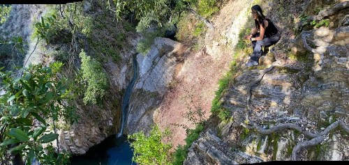 A person sits on a rocky ledge overlooking a lush canyon with a waterfall and clear water below.