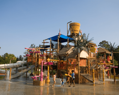 A colorful water play structure with slides and water features, surrounded by trees and families enjoying the splash area.