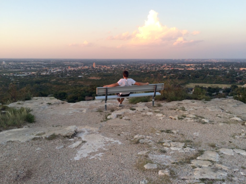 A person sits on a bench overlooking a scenic landscape at sunset, with a vast view of the city below.