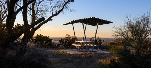 A picnic table under a shelter at sunset, surrounded by trees and open landscape.