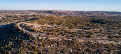 Aerial view of a hilly landscape with sparse vegetation, roads, and distant wind turbines under a clear sky.