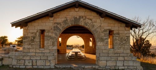 A stone pavilion with an arched entrance, featuring picnic tables inside, set against a sunset backdrop.