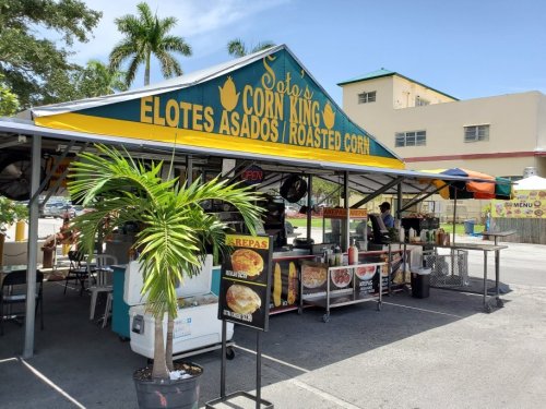 A colorful food stand named "Soto's Corn King" selling elotes asados and roasted corn, with palm trees in the background.