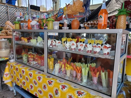 A colorful display of fresh fruit cups, desserts, and drinks in a market stall, decorated with vibrant items.