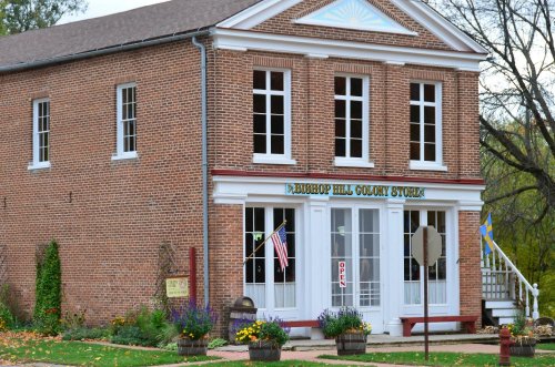 Historic brick building with "Bishop Hill Colony Store" sign, open flag, and colorful flower planters outside.