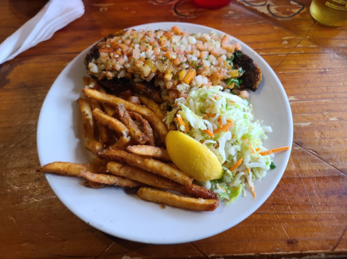 A plate of grilled fish topped with diced tomatoes and onions, served with fries, coleslaw, and a lemon wedge.