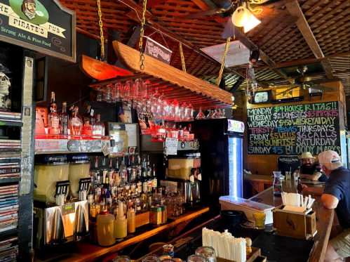 A lively bar scene featuring a wooden boat overhead, shelves of liquor, and a chalkboard menu with drink specials.